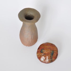 May include: A small, brown, textured clay vase with a wide, flared rim and a separate, smooth, circular lid featuring a polished, multi-colored surface with streaks of red, orange, and dark blue, presented together on a plain white background.