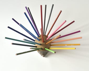 Puede incluir: Un portalápices de cerámica marrón con forma de erizo y superficie texturizada, que presenta numerosos orificios en el lomo. Varios lápices de colores en tonos azul, verde, amarillo, rosa, morado, marrón y negro están insertados.