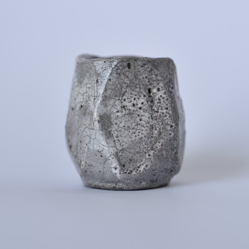 May include: A small, gray ceramic cup with a textured, crackled glaze. The cup has a rounded shape and a slightly flared rim.
