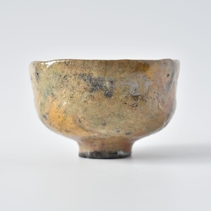 May include: A small, round ceramic bowl with a textured, uneven surface. The exterior features a mottled glaze in shades of tan, ochre, and muted brown, with dark grey and black speckles and patches throughout the finish. The bowl has a thick rim.