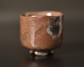 Puede incluir: Una pequeña taza de cerámica cilíndrica con una superficie esmaltada de color marrón rojizo que presenta un patrón de grietas finas. El exterior muestra variaciones sutiles de color, incluyendo manchas gris oscuro y blancas, con un borde grueso e irregular.