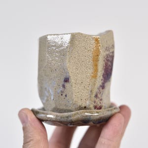 May include: A handmade ceramic planter with a matching saucer. The planter has a geometric, faceted design with a neutral beige color, accented by streaks of purple and gold. The saucer has a wavy edge.