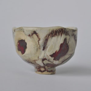 May include: A small, white ceramic bowl with a textured, uneven surface. The bowl has a red and brown glaze with a mottled pattern. The bowl is sitting on a white surface.
