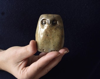 Puede incluir: Una pequeña figura de búho de cerámica en color beige, con una superficie lisa y acabado brillante. El búho presenta dos ojos oscuros, circulares y hundidos, junto con marcas incisas en la base que representan las patas, forma compacta.