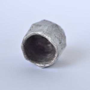 May include: A small, gray ceramic cup with a textured, cracked glaze. The cup has a hexagonal shape and a wide, open mouth.