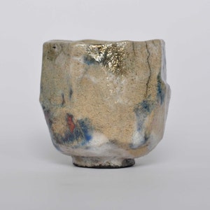 May include: A ceramic cup with a textured, uneven surface featuring a beige base color. The exterior is decorated with patches of blue and red pigment, along with metallic gold accents. The rim is irregular and the base is dark and narrow.