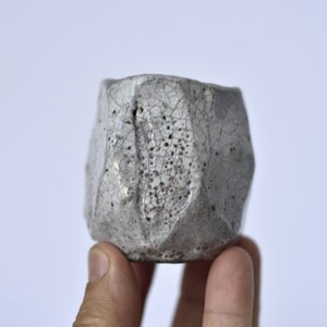 May include: A small, white ceramic cup with a textured, crackled glaze. The cup has a geometric shape with many sides.