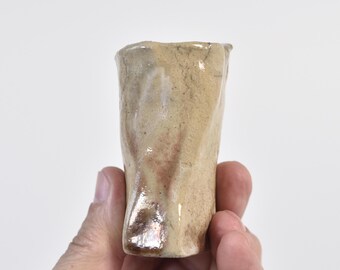 Puede incluir: Una pequeña taza de cerámica hecha a mano con una forma orgánica e irregular. La superficie presenta un esmalte moteado en tonos beige y bronceado con sutiles reflejos marrones y naranja rojizo, acabado brillante y borde irregular.
