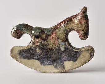 Puede incluir: Una figura de caballo balancín de cerámica con un esmalte brillante y moteado en tonos marrón tierra, verde salvia y carbón oscuro. La superficie tiene un aspecto texturizado e irregular con un acabado reflectante que destaca los colores.