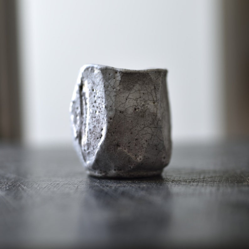 May include: A small, gray ceramic cup with a textured, cracked surface. The cup is slightly tilted to the side, revealing its irregular shape.