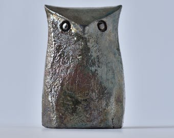 Puede incluir: Una figura de búho estilizada con un acabado metálico texturizado en tonos gris oscuro, bronce y azul apagado. El objeto presenta una parte superior puntiaguda, una base lisa y plana, y dos pequeños ojos circulares de borde oscuro en la parte superior.