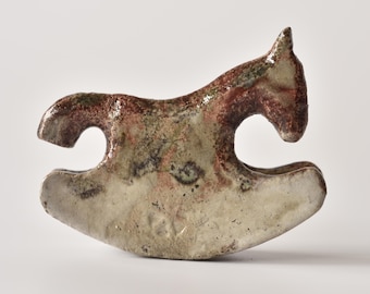 Puede incluir: Una figura de caballo balancín de cerámica con un esmalte brillante y moteado en tonos crema, marrón y verde apagado. La escultura tiene una forma estilizada y abstracta con una base curva, una cabeza definida y una textura superficial lisa.