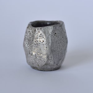 May include: A small, gray ceramic cup with a textured, crackled glaze. The cup has a rounded, slightly irregular shape.