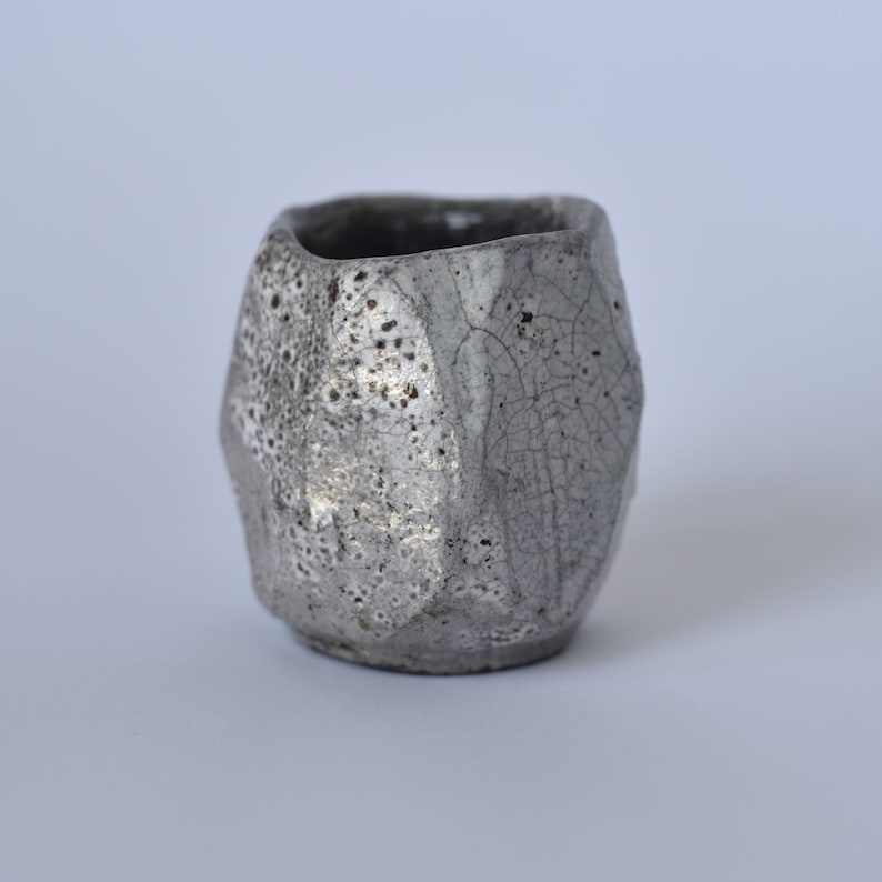 May include: A small, gray ceramic cup with a textured, crackled glaze. The cup has a rounded, slightly irregular shape.