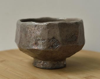 Puede incluir: Un pequeño cuenco de cerámica formado a mano con una superficie exterior facetada e irregular. El cuenco presenta un esmalte moteado en tonos tierra marrones y grises con un acabado texturizado e irregular y una base circular elevada.