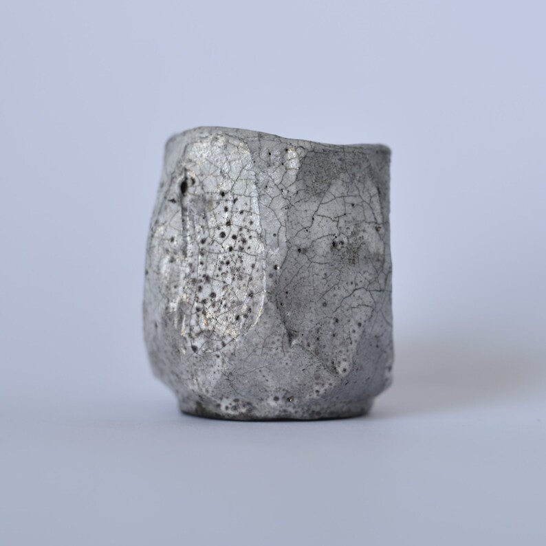 May include: A small, silver ceramic cup with a textured, cracked surface. The cup has a rounded shape and a slightly uneven rim.