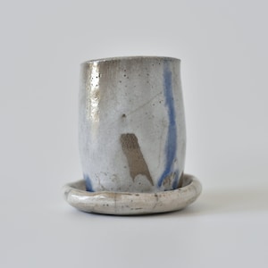 Könnte beinhalten: Eine weiße Keramiktasse mit blauem und braunem Glase, die auf einer passenden Untertasse steht. Tasse und Untertasse haben ein rustikales, handgemachtes Aussehen.