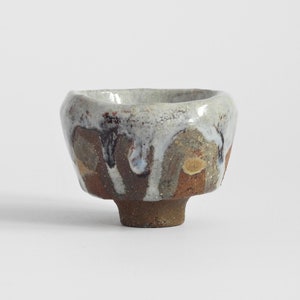 May include: A small, hand-thrown ceramic tea bowl with a thick, uneven white glaze dripping over a textured, earthy brown and tan clay body. The vessel features a wide, flared rim and a sturdy, cylindrical foot, showcasing a rustic finish.