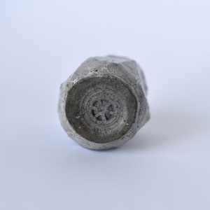 May include: A gray ceramic vase with a rough, textured surface. The vase has a circular opening at the bottom with a decorative design.