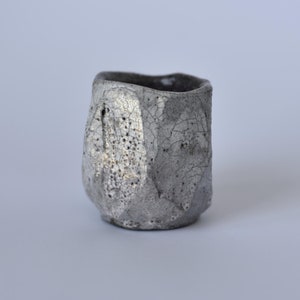 May include: A small, gray ceramic cup with a textured, crackled glaze. The cup has a slightly irregular shape and a wide, rounded base.