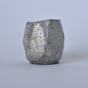 May include: A small, gray ceramic cup with a textured, crackled glaze. The cup has a unique, geometric shape.