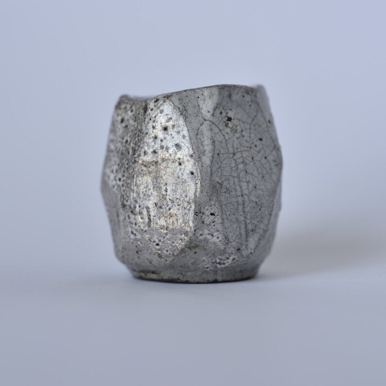 May include: A small, gray ceramic cup with a textured, crackled glaze. The cup has a unique, geometric shape.