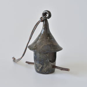 May include: A small, dark, glazed ceramic birdhouse with a conical roof and a circular opening. A thin, natural wooden twig is inserted horizontally through the base to serve as a perch. A grey and orange braided cord is attached to the top loop.