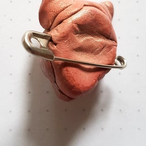 Puede incluir: Una peque&ntilde;a escultura de arcilla rosa de la cabeza de un perro con un imperdible sujeto a la parte posterior.