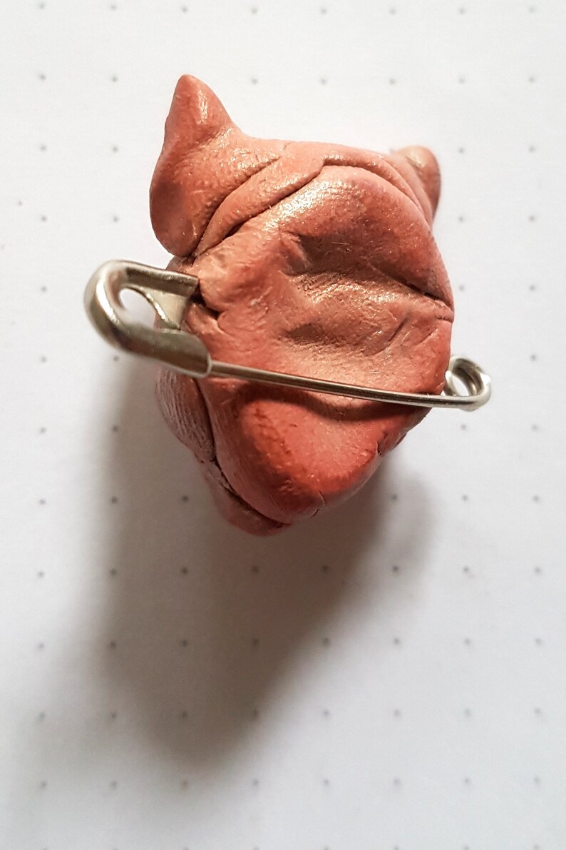 Puede incluir: Una peque&ntilde;a escultura de arcilla rosa de la cabeza de un perro con un imperdible sujeto a la parte posterior.