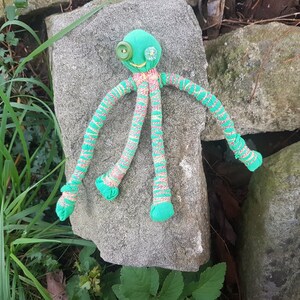 May include: A green fabric octopus with button eyes and striped legs. The octopus is lying on a gray rock.