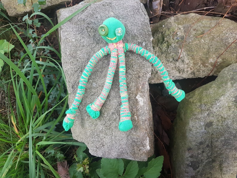 May include: A green fabric octopus with button eyes and striped legs. The octopus is lying on a gray rock.