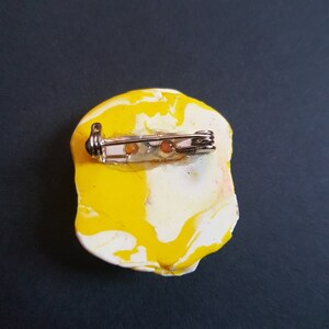 May include: A yellow and white marbled brooch with a silver pin and clasp. The brooch is shaped like a rounded rectangle.