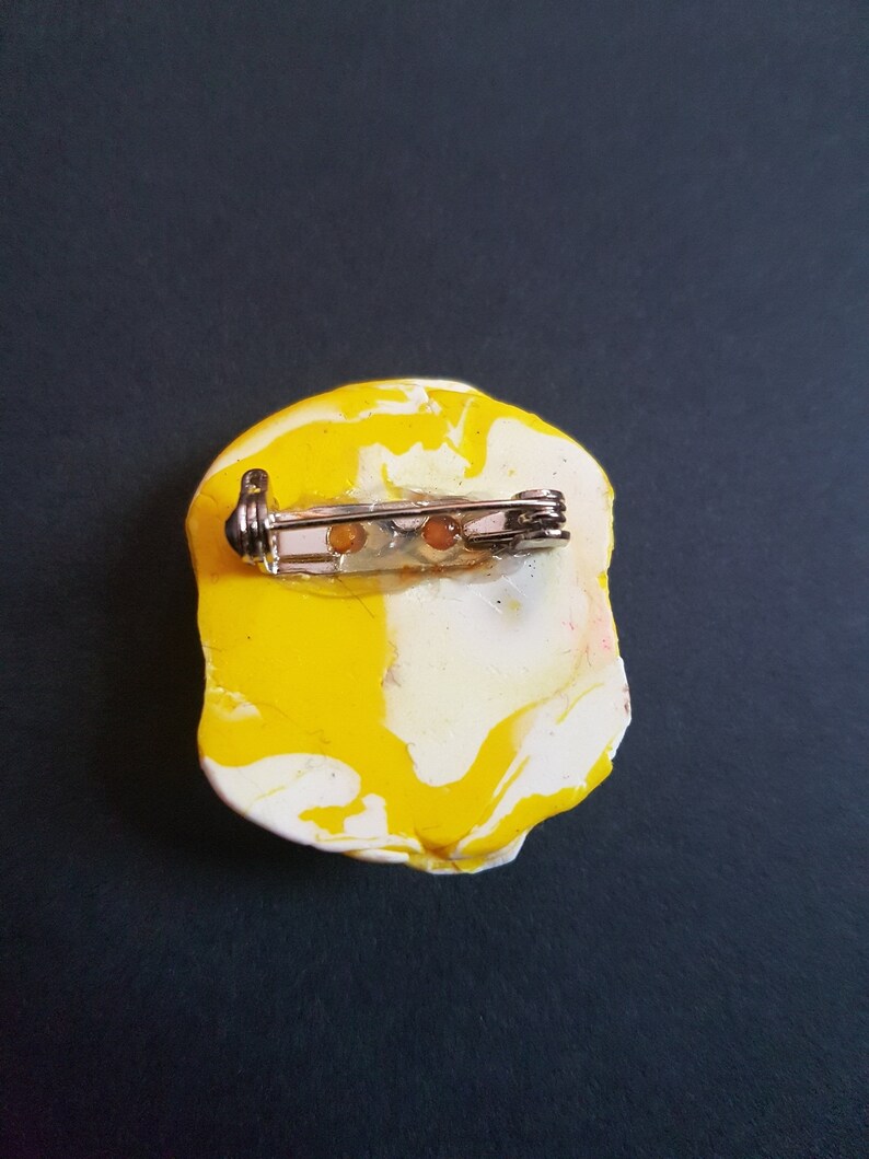 May include: A yellow and white marbled brooch with a silver pin and clasp. The brooch is shaped like a rounded rectangle.