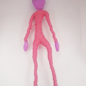 May include: A pink yarn doll with a purple head and white feet. The doll is standing on a white background.