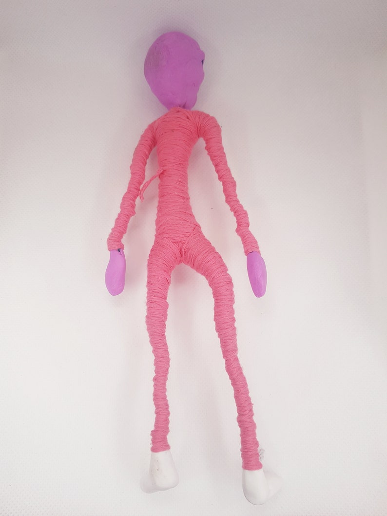 May include: A pink yarn doll with a purple head and white feet. The doll is standing on a white background.