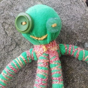 May include: A green fabric doll with red and white striped arms and legs. The doll has two buttons for eyes, one green and one blue.