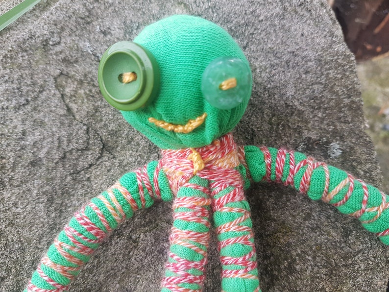May include: A green fabric doll with red and white striped arms and legs. The doll has two buttons for eyes, one green and one blue.