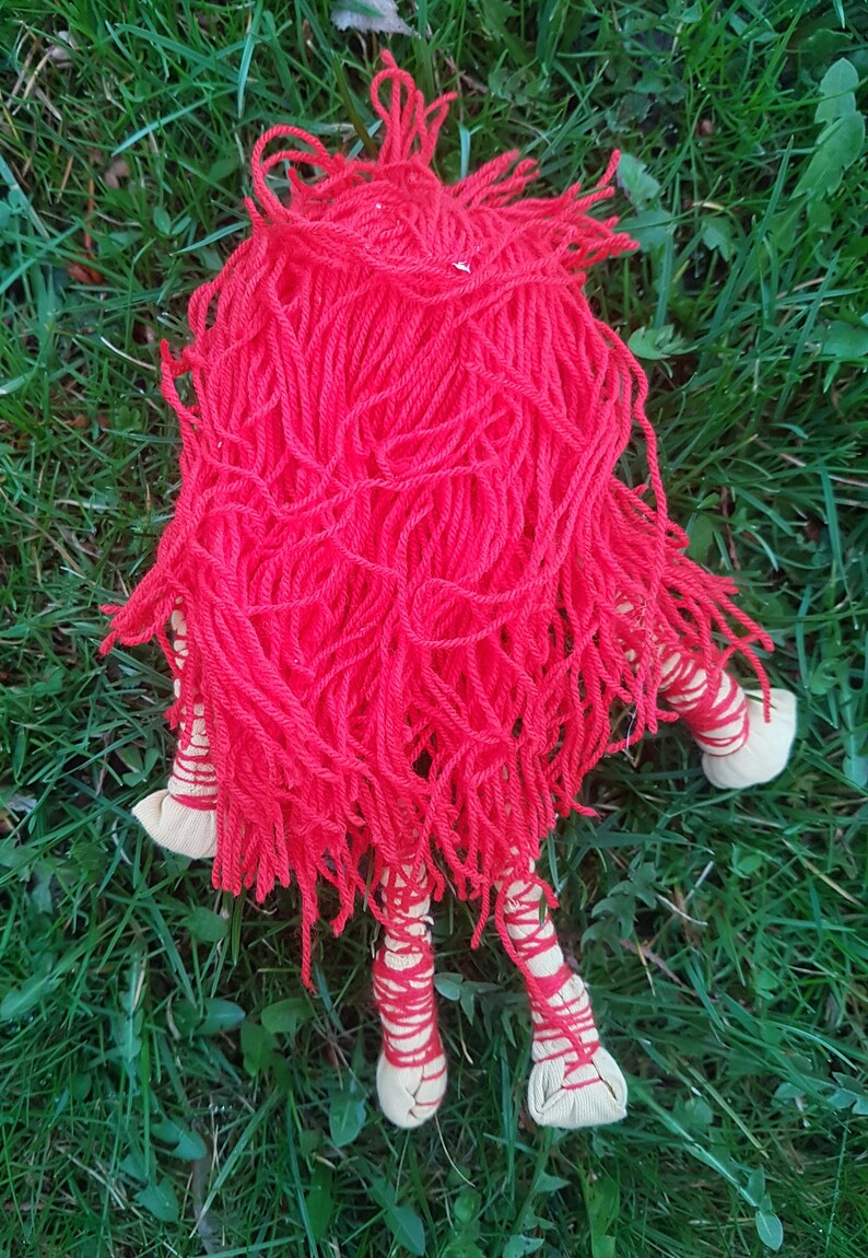 May include: A handmade doll with a vibrant red yarn head and body. The doll's legs are wrapped in red yarn and have beige fabric feet. The doll is lying on a bed of green grass.