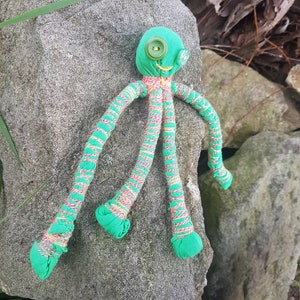May include: A green fabric octopus with striped legs and button eyes. The octopus is lying on a gray rock.