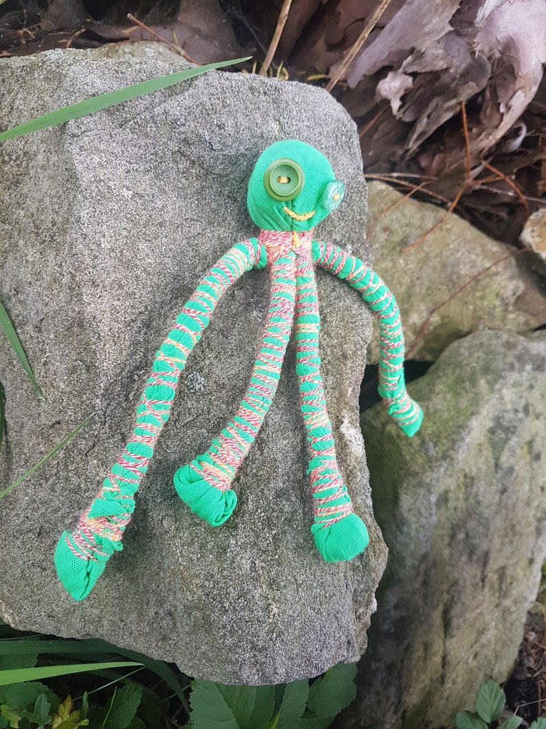 May include: A green fabric octopus with striped legs and button eyes. The octopus is lying on a gray rock.