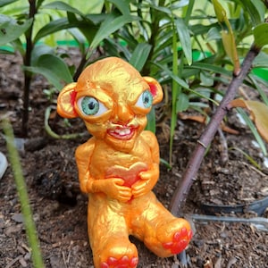 May include: A whimsical, orange-gold figurine of a seated creature with large eyes and a wide grin, holding a heart. The figurine has red accents on its feet and ears. The sculpture is set against a backdrop of green foliage and brown soil.
