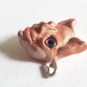 Puede incluir: Una escultura de arcilla rosa y marr&oacute;n de una cara grotesca con un ojo morado y verde. La escultura est&aacute; unida a un pasador de seguridad plateado.