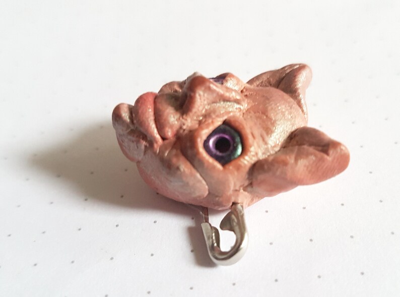 Puede incluir: Una escultura de arcilla rosa y marr&oacute;n de una cara grotesca con un ojo morado y verde. La escultura est&aacute; unida a un pasador de seguridad plateado.