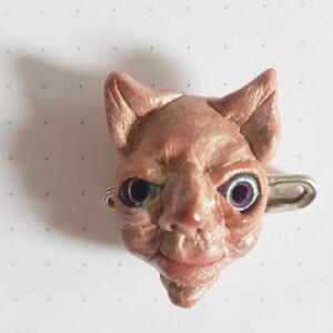 Puede incluir: Una peque&ntilde;a escultura de arcilla rosa de una cabeza de gato con ojos verdes y un pasador de seguridad plateado sujeto a la parte posterior. La escultura est&aacute; detallada con arrugas y una leve sonrisa.