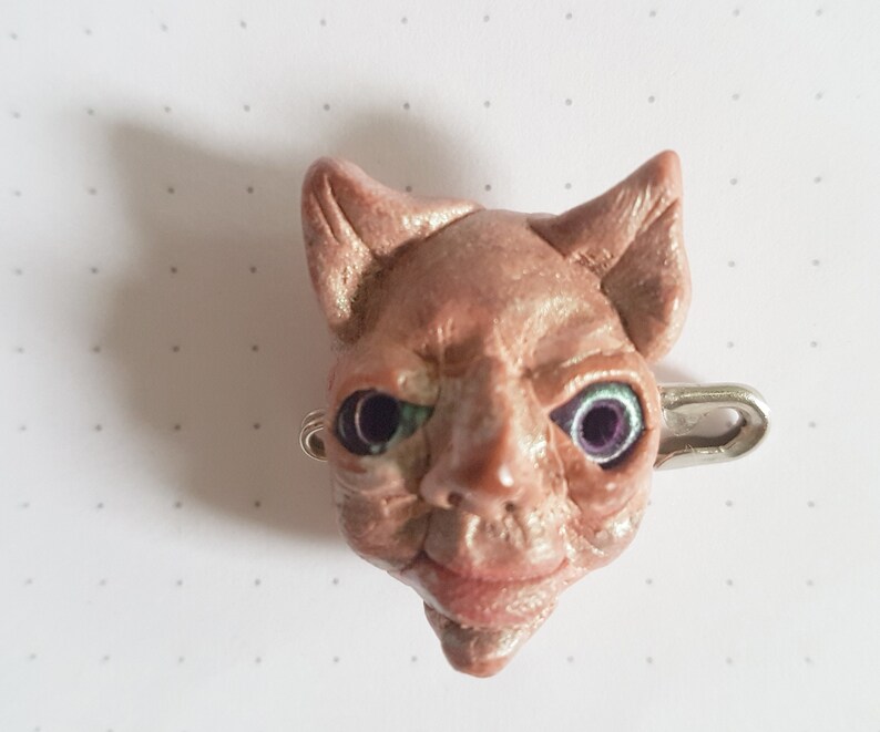 Puede incluir: Una peque&ntilde;a escultura de arcilla rosa de una cabeza de gato con ojos verdes y un pasador de seguridad plateado sujeto a la parte posterior. La escultura est&aacute; detallada con arrugas y una leve sonrisa.