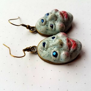 May include: A pair of earrings featuring a whimsical, textured, white face with blue eyes and red lips. The earrings are made of polymer clay and have a bronze-coloured metal hook.