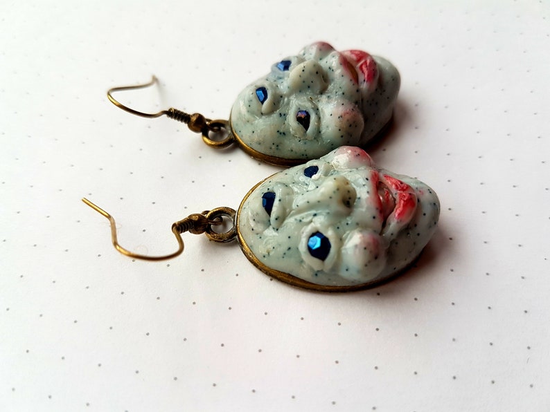 May include: A pair of earrings featuring a whimsical, textured, white face with blue eyes and red lips. The earrings are made of polymer clay and have a bronze-coloured metal hook.