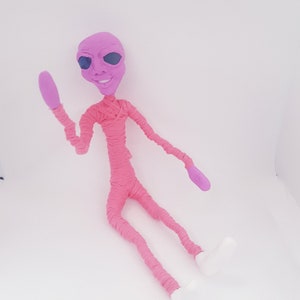 May include: A pink alien doll with large eyes and a smile, made of yarn and clay. The doll is sitting with one arm raised in a wave.