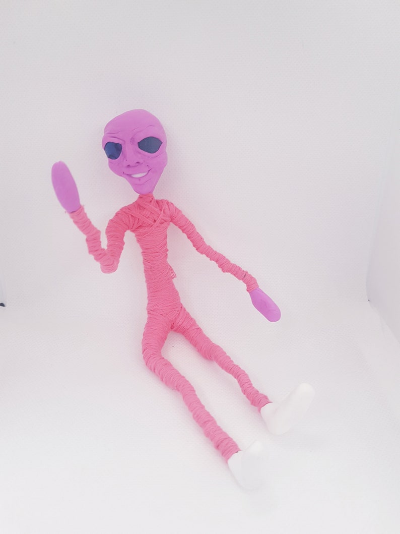 May include: A pink alien doll with large eyes and a smile, made of yarn and clay. The doll is sitting with one arm raised in a wave.