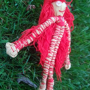 May include: Handmade doll with a fabric face, button eyes, and red yarn hair. The doll's arms and legs are wrapped in cream-colored fabric and red yarn. The doll is lying on green grass.